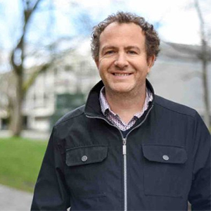 A smiling Lucas Wright with curly brown hair, wearing a dark jacket over a patterned collared shirt, in an outdoor setting.